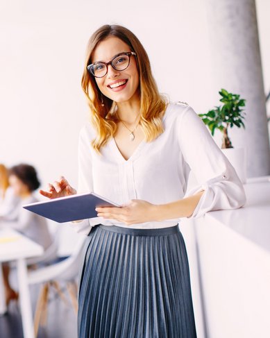 Eine Frau mit Brille hält ein Tablet und lächelt, während sie in einem modernen Büro steht. Im Hintergrund sind mehrere Personen an einem Tisch mit Laptops und Dokumenten beschäftigt.