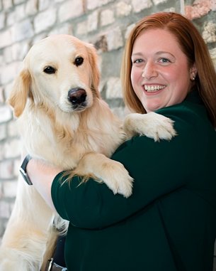Eine Frau mit langen, roten Haaren hält einen Golden Retriever in ihren Armen. Sie lächelt und steht vor einer gemauerten Wand.
