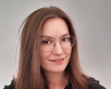 Eine Person mit langen, glatten Haaren und einer schwarzen Bluse trägt runde Brille und lächelt leicht in die Kamera. Der Hintergrund ist neutral und hell.