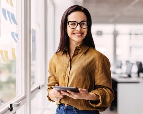 Eine Frau mit Brille steht in einem modernen Büro und hält ein Tablet in der Hand. Sie trägt ein braunes Hemd und lächelt, während sie an einem Fenster steht, das mit bunten Notizen dekoriert ist.
