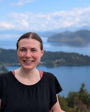 Eine Frau mit langen Haaren lächelt vor einer malerischen Seenlandschaft mit Bergen im Hintergrund.