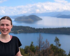 Eine Frau mit langen Haaren lächelt vor einer malerischen Seenlandschaft mit Bergen im Hintergrund.