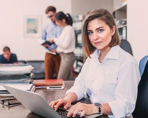 Eine Frau sitzt an einem Schreibtisch und arbeitet an einem Laptop. Im Hintergrund sind zwei Personen zu sehen, die miteinander sprechen, während ein weiterer Mann auf einem Stuhl sitzt und liest.