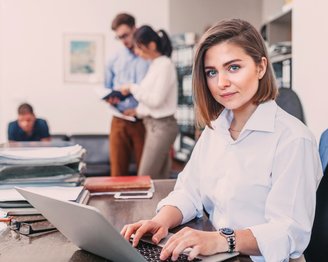 Eine Frau sitzt an einem Schreibtisch und arbeitet an einem Laptop. Im Hintergrund sind zwei Personen zu sehen, die miteinander sprechen, während ein weiterer Mann auf einem Stuhl sitzt und liest.