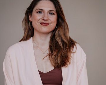 Eine Frau mit langen, lockigen Haaren trägt ein rosa Oberteil und einen braunen BH. Sie steht vor einem neutralen Hintergrund und lächelt leicht.