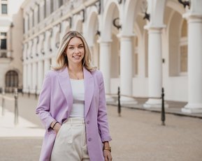 Eine Frau mit langen, blonden Haaren trägt einen lila Blazer und eine weiße Bluse. Sie steht in einer architektonisch gestalteten Umgebung mit Säulen und einem gepflasterten Boden.