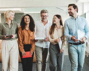 Fünf Personen in formeller Kleidung gehen lächelnd durch ein modernes Büro. Sie halten Notizen und Tablets in den Händen und scheinen sich angeregt zu unterhalten.