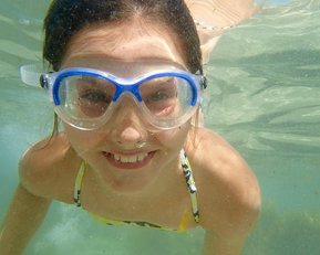 Ein Mädchen mit Schwimmbrille taucht unter Wasser und lächelt. Sie trägt einen bunten Badeanzug und ist umgeben von klarem Wasser.