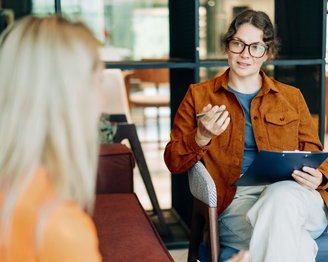 Berufe in der klinischen Psychologie ohne Approbation Eine Frau mit Brille und braunem Blazer sitzt auf einem Stuhl und spricht mit einer anderen Person, während sie ein Notizblatt hält.