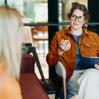 Eine Frau mit Brille und braunem Blazer sitzt auf einem Stuhl und spricht mit einer anderen Person, während sie ein Notizblatt hält.