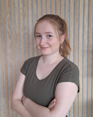 Junge Frau mit blonden Haaren, die in einem grünen Oberteil gekleidet ist, steht mit verschränkten Armen vor einer gestreiften Wand.
