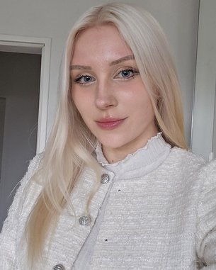 Eine Person mit langen, blonden Haaren trägt eine helle Jacke und schaut in die Kamera. Der Hintergrund zeigt einen neutralen Raum mit einer Tür.