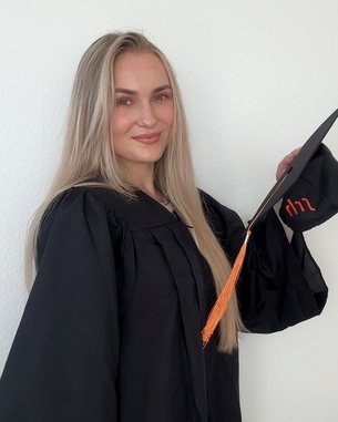 Eine junge Frau in einem schwarzen Abschlussmantel hält einen Doktorhut mit einer orangefarbenen Quaste. Sie lächelt und steht vor einer neutralen Wand.