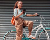 Eine junge Frau sitzt auf einem weißen Fahrrad, hält ein Smartphone in der Hand und lächelt. Sie trägt eine hellblaue Bluse, beige Hosen und weiße Sneakers. Im Hintergrund sind graue Rolltore sichtbar.